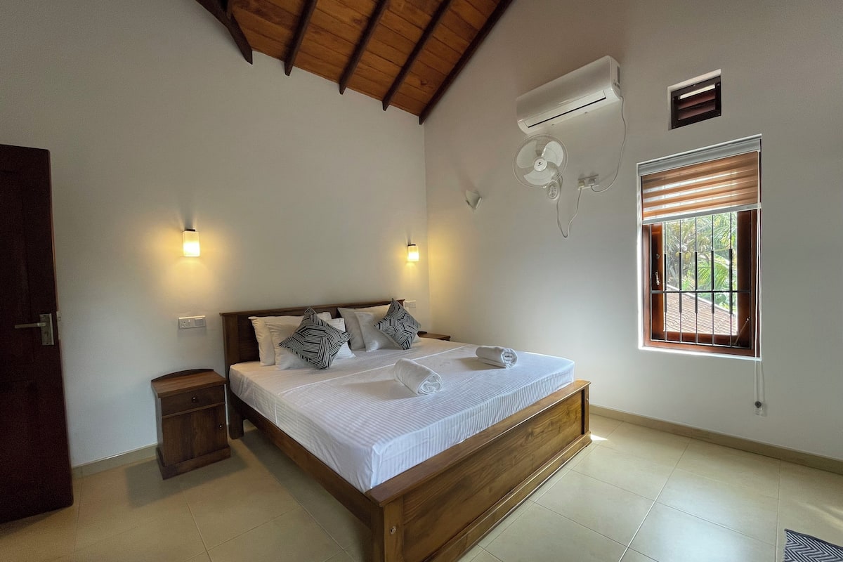 A bedroom with a wooden bed, two bedside tables, an air conditioning unit, a window with blinds, and a wall-mounted fan.