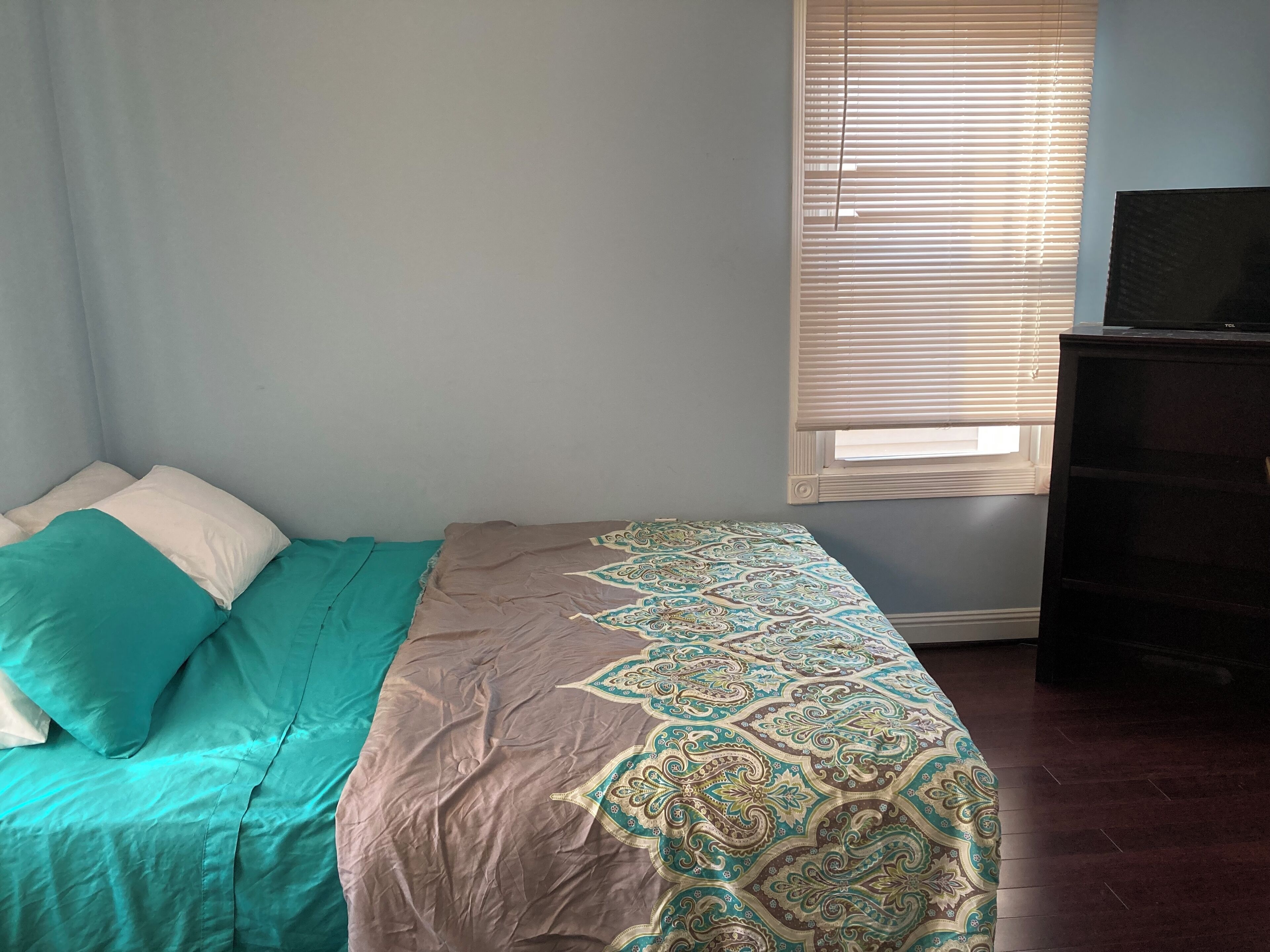 A bedroom with a bed, a window with blinds, and a dark dresser.