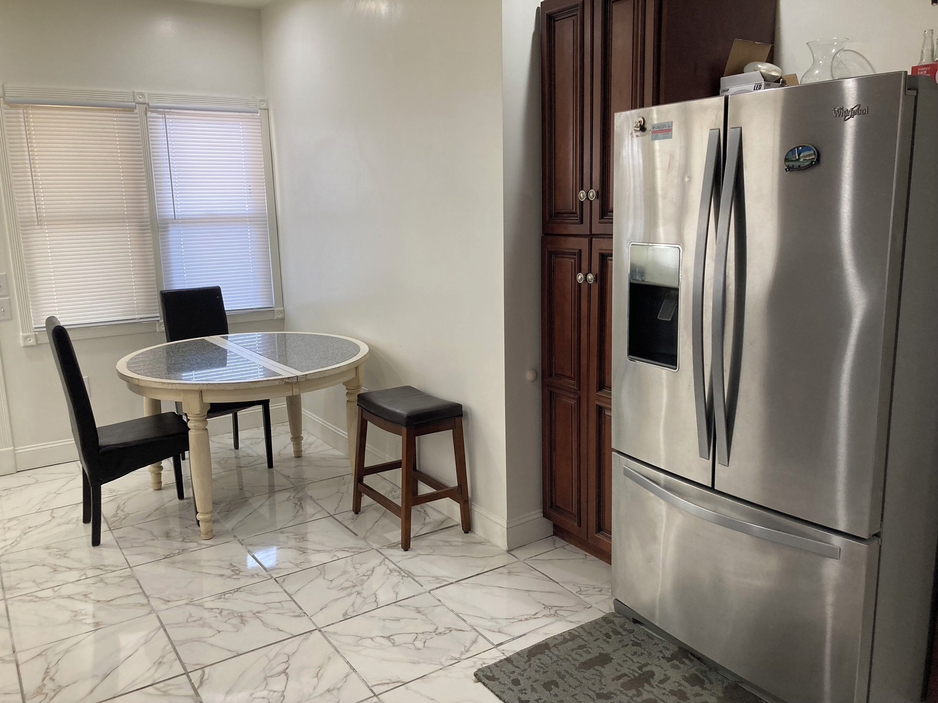A kitchen with a marble floor, a stainless steel refrigerator, a dining table with chairs, and a window with blinds.