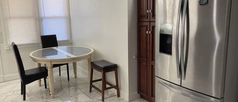 A kitchen with a marble floor, a stainless steel refrigerator, a dining table with chairs, and a window with blinds.