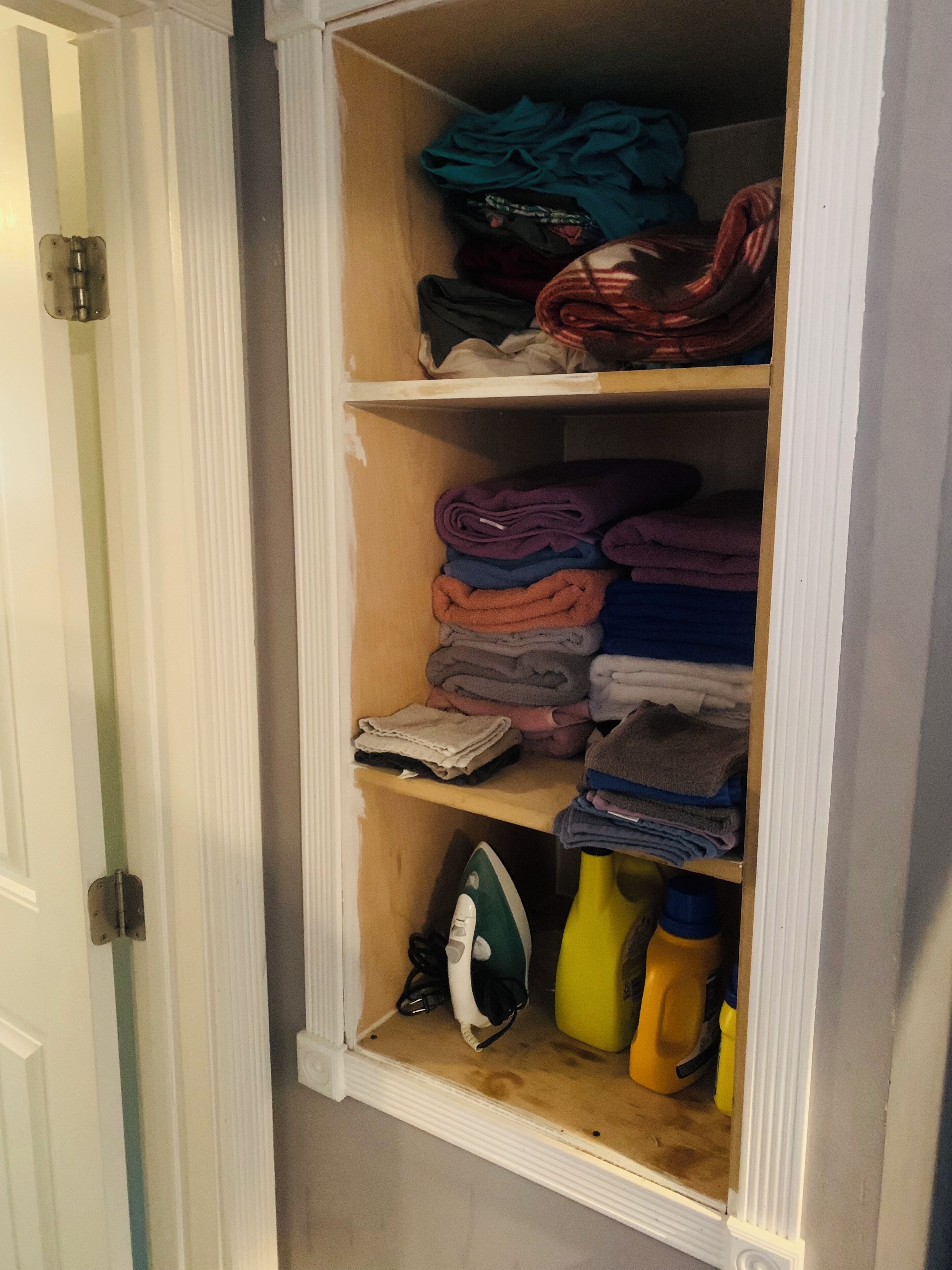 A closet with shelves holding folded clothes, an ironing board, and cleaning supplies.