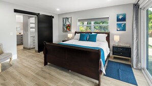 A bedroom with a large bed, a nightstand, a window with a view of greenery, and a sliding door leading to a bathroom. - Inspiration House-One Acre Pool Home in the Heart of Pinellas!  (Pinellas Park)