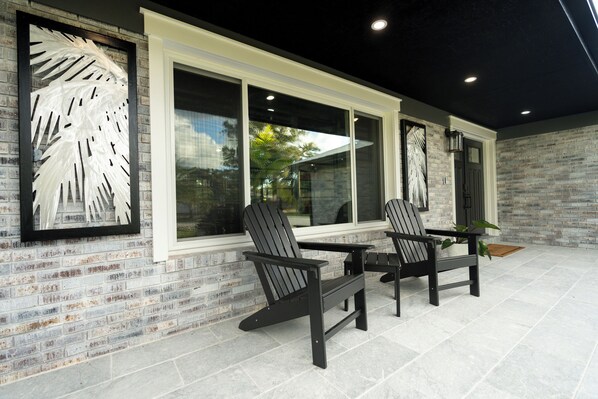 An outdoor patio with two Adirondack chairs, a table, and a brick wall with large windows. - Inspiration House-One Acre Pool Home in the Heart of Pinellas! (Pinellas Park)