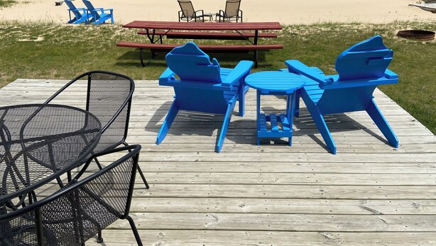 Una terraza de madera con sillas Adirondack azules y una mesa de metal, con vista a una playa que tiene una mesa de picnic y sillas de descanso.