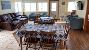 Una sala de estar con un sofá de cuero marrón, una mesa de comedor con mantel de tela estampada en azul, sillas de madera, un sillón azul y un televisor en la pared.