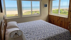 Un dormitorio con una cama, una lámpara y tres ventanas con vistas a la playa.