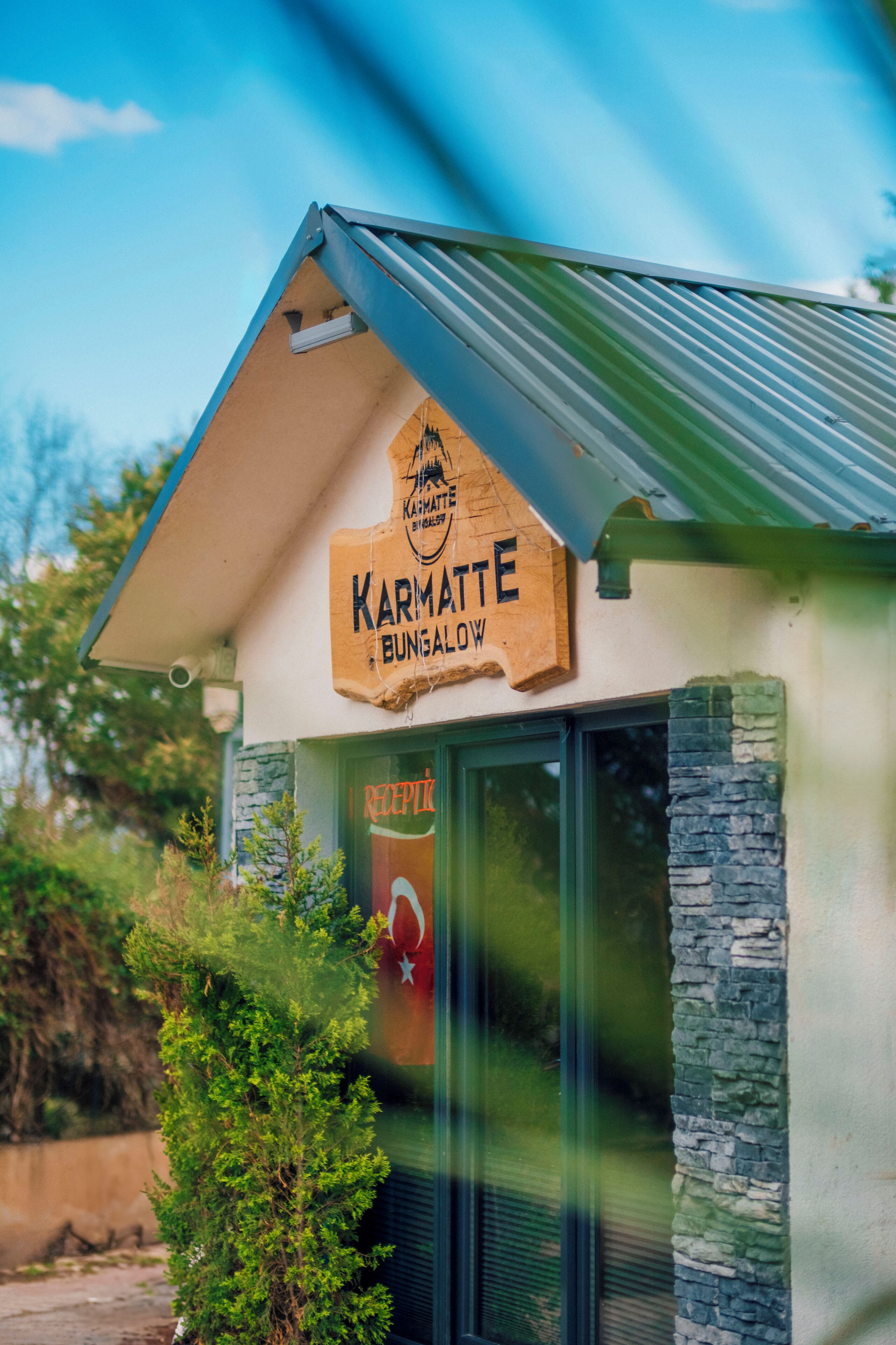 Ein Bungalow mit einem Schild, auf dem 'Karmatte Bungalow' steht, und einem durch das Fenster einsehbarer Rezeptionsbereich.