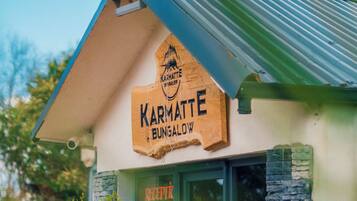 Ein Bungalow mit einem Schild, auf dem 'Karmatte Bungalow' steht, und einem durch das Fenster einsehbarer Rezeptionsbereich.