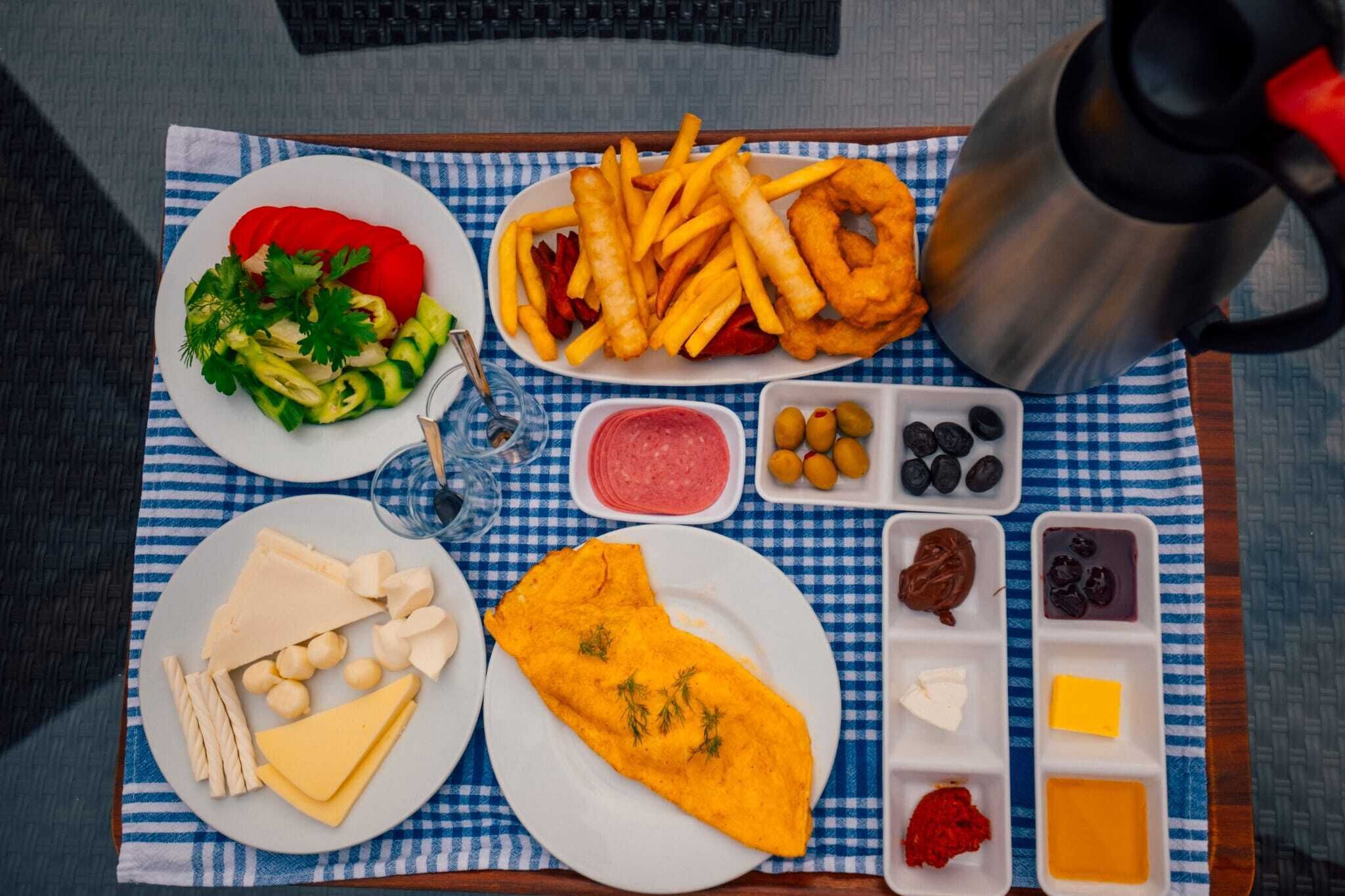 Uma bandeja de refeição com diversos alimentos, incluindo batatas fritas, queijo e uma omelete, acompanhada de acompanhamentos e azeitonas.