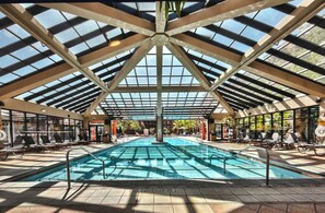 A large indoor swimming pool with a glass ceiling, surrounded by lounge chairs and a sunlit poolside area.