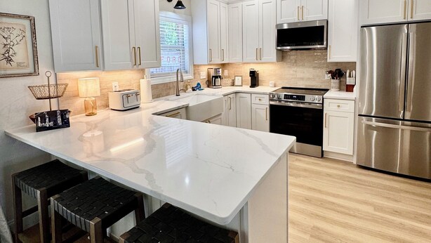 A modern kitchen with white cabinetry, a large island with bar stools, stainless steel appliances, and a window with blinds. -     Freshly Remodeled Oxford Retreat 1 Mile to Ole Miss & The Square (Oxford)