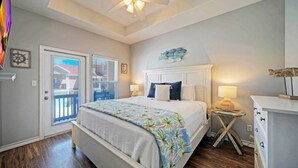 A bedroom with a bed, a ceiling fan, a window with blinds, a door, and a small table with a lamp. - Walk to Beach Shared Pool Access, Golf Cart Zone Carpe Beach Bum by Avantstay (Port Aransas)