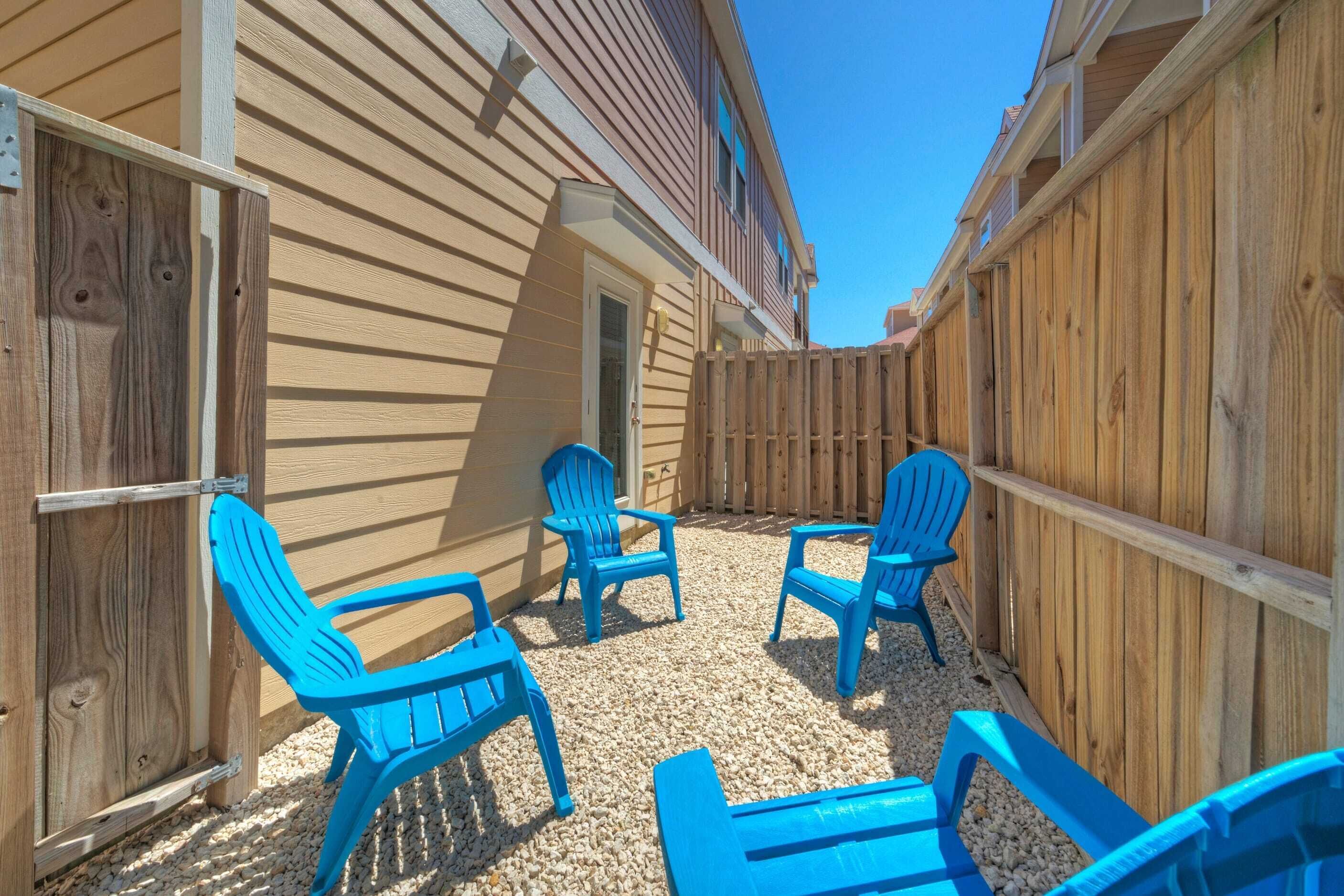 A covered outdoor patio with four blue Adirondack chairs and a small table, surrounded by wooden fencing and a building with beige siding.