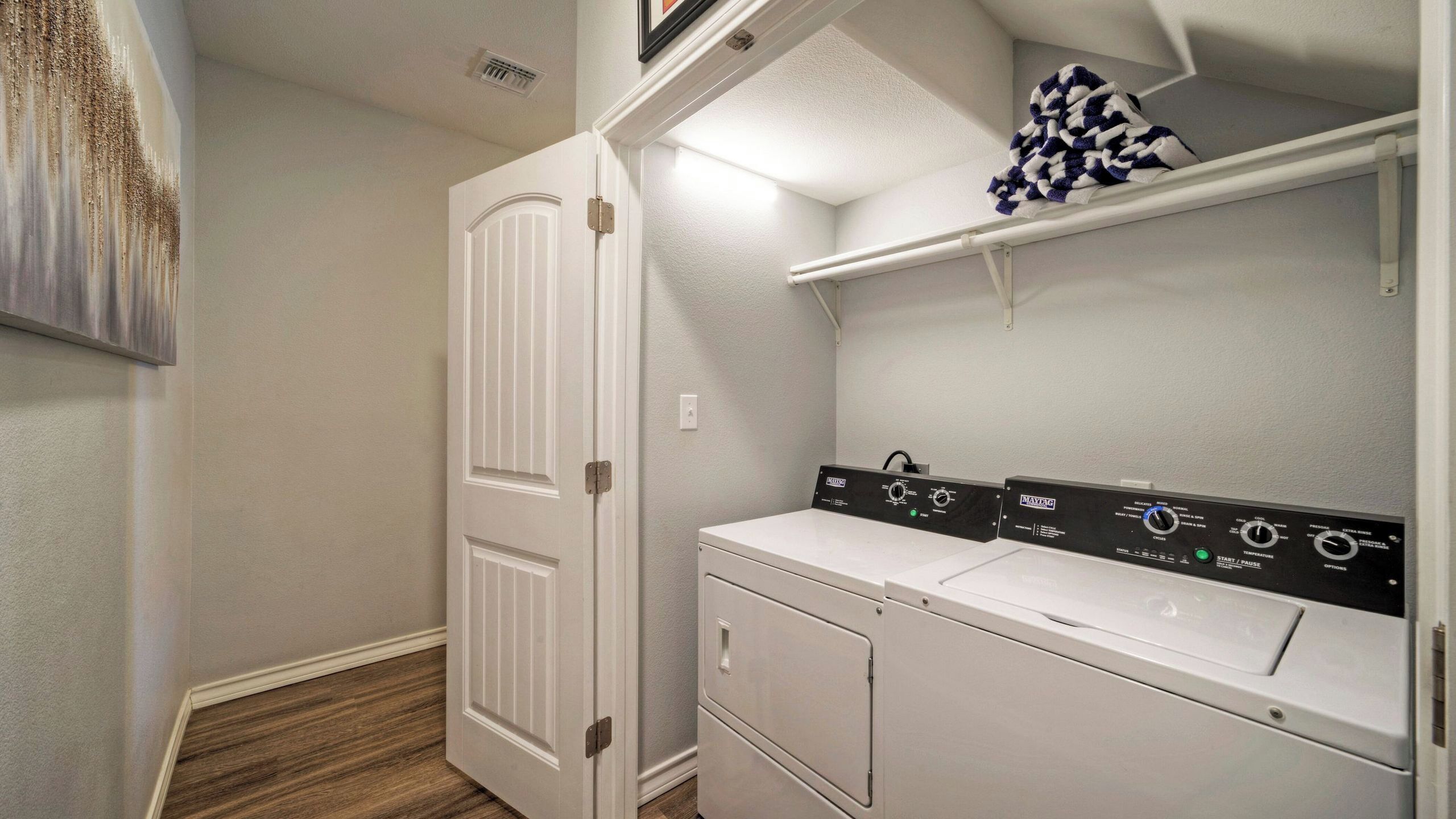A laundry room with a white door, a washing machine, and a dryer. There are control panels on the dryer and a shelf above it.
