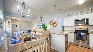 A modern kitchen with white cabinets, a built-in microwave, and an oven. There is a dining area with a wooden table and chairs, and a TV on the wall. - Walk to Beach Shared Pool Access, Golf Cart Zone Carpe Beach Bum by Avantstay (Port Aransas)