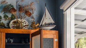 Een kamer met een houten kastje, daarin keukengerei, een decoratie van een zeilboot en een raam met uitzicht op bomen.