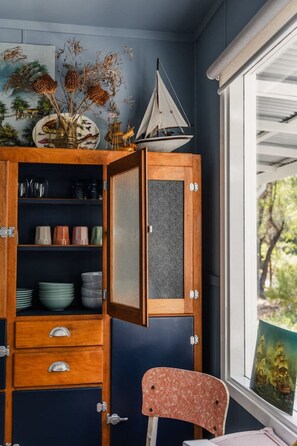 A room with a wooden cabinet containing kitchenware, a sailboat decoration, and a window with a view of trees. - Poppy`s Place - Denmark (Denmark)