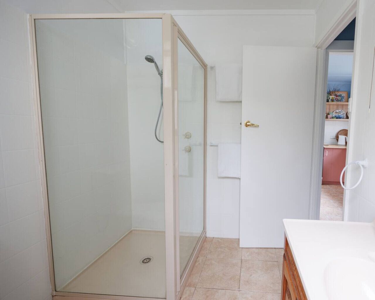 A bathroom with a glass-enclosed shower, a white sink, and a door leading to a kitchen area.