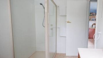 A bathroom with a glass-enclosed shower, a white sink, and a door leading to a kitchen area.