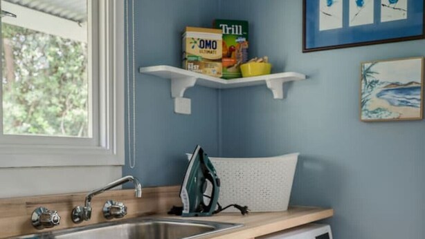 Een wasserette met een wasmachine, een spoelbak en een raam. Boven de spoelbak staan planken met verschillende spullen.