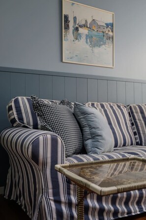 A striped sofa with a checkered pillow and a woven coffee table against a wall with a framed painting.