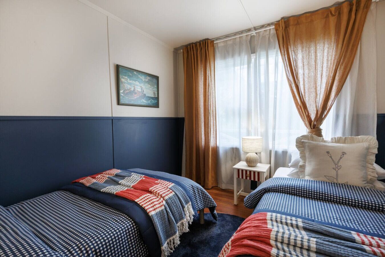 A hotel room with two beds, a nightstand with a lamp, a painting on the wall, and a window with curtains.