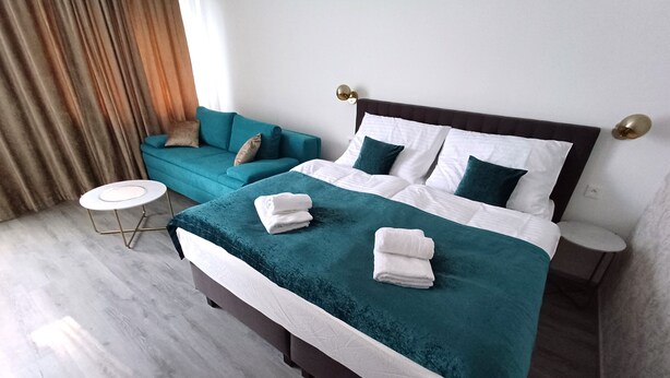 A neatly made bed with a teal bedspread and white linens, a small round table with a gold base, and a teal sofa with a brown cushion. - LUNEX Apartment (Jihočeský kraj)