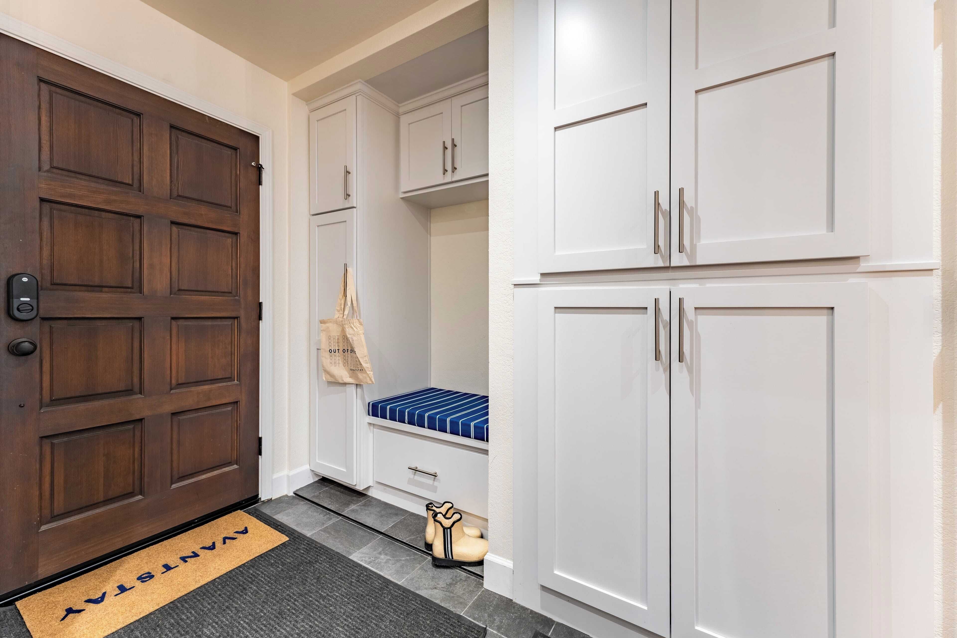 Un couloir avec une porte en bois, des armoires blanches, un banc doté d’un coussin à rayures bleues et blanches, et un petit objet décoratif posé sur le sol.