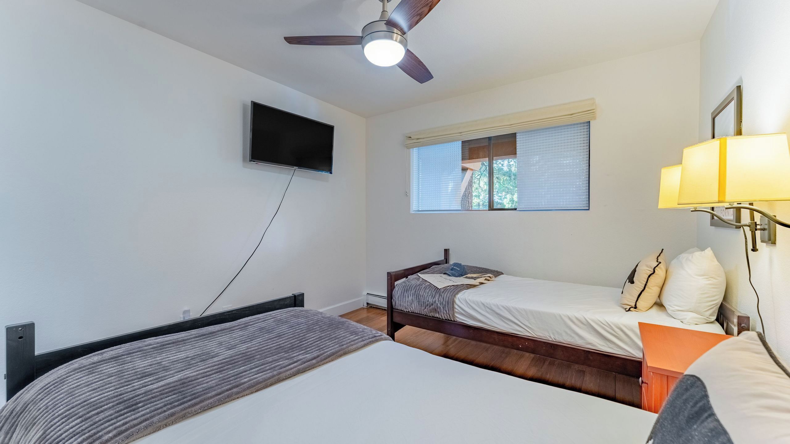 Habitación con dos camas, ventilador de techo, televisor montado en la pared y ventana con persianas.