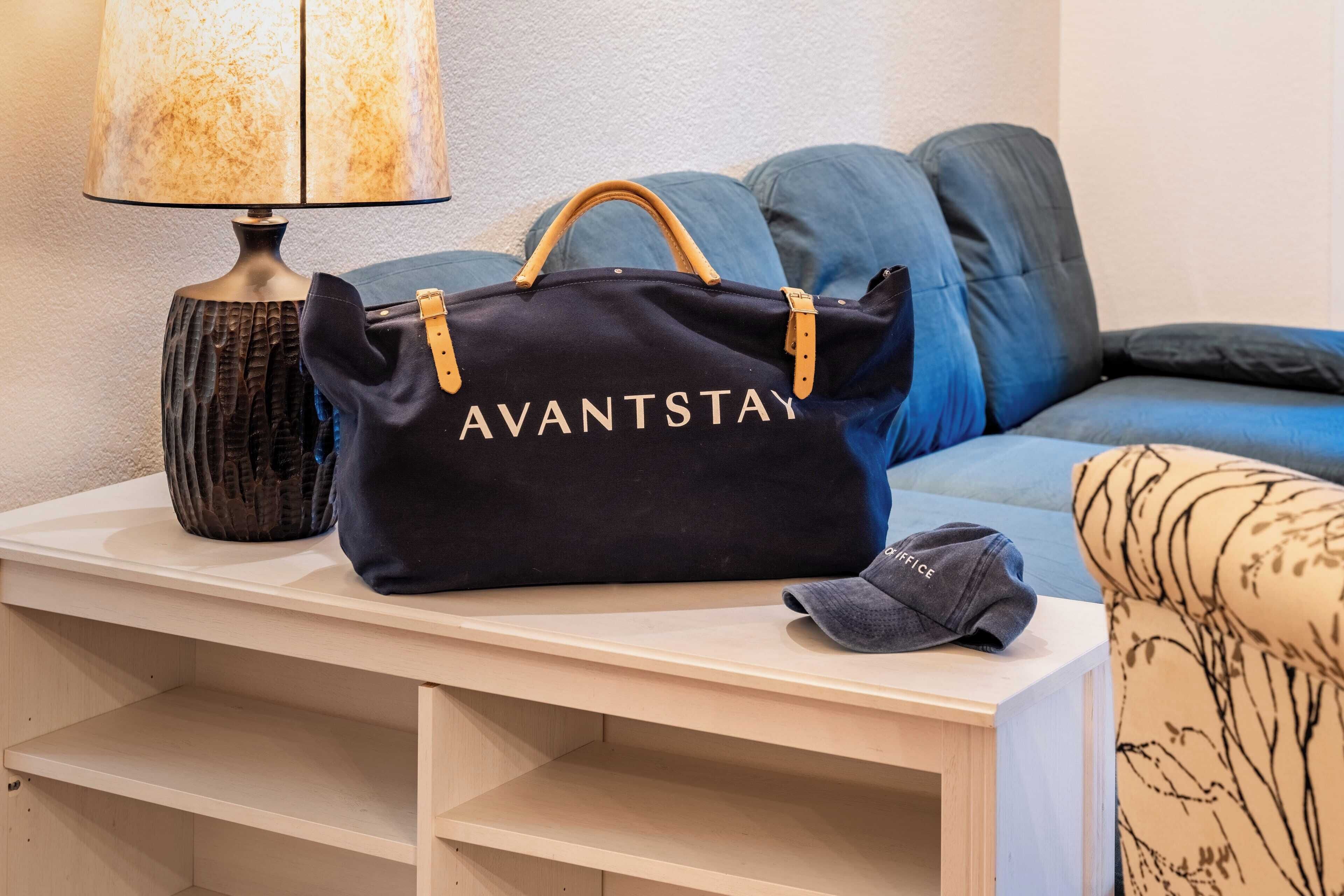 Un sac noir avec l’inscription « AVANTSTAY », une lampe et un canapé avec des coussins bleus.