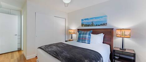 A bedroom with a bed, a ceiling fan, a nightstand with a lamp, and a painting on the wall.