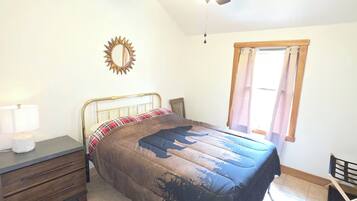 Een slaapkamer met een bed, een plafondventilator, een raam met gordijnen, een nachtkastje en een stoel.