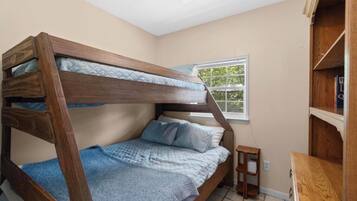 A bunk bed with a wooden frame and blue bedding in a room with a window and a small wooden side table.