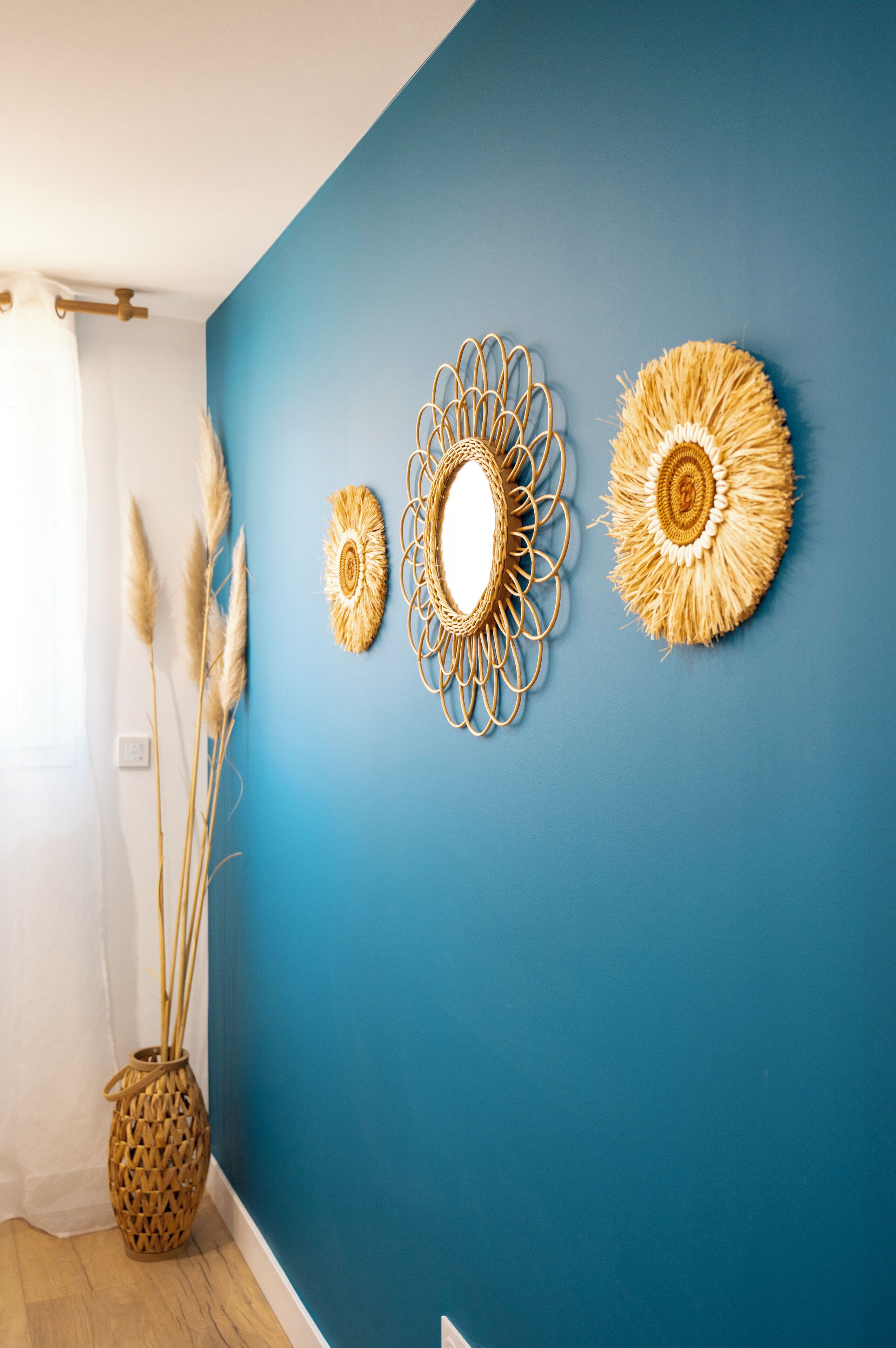 Une pièce avec un mur bleu, trois éléments décoratifs suspendus au mur et un vase en osier contenant des roseaux.