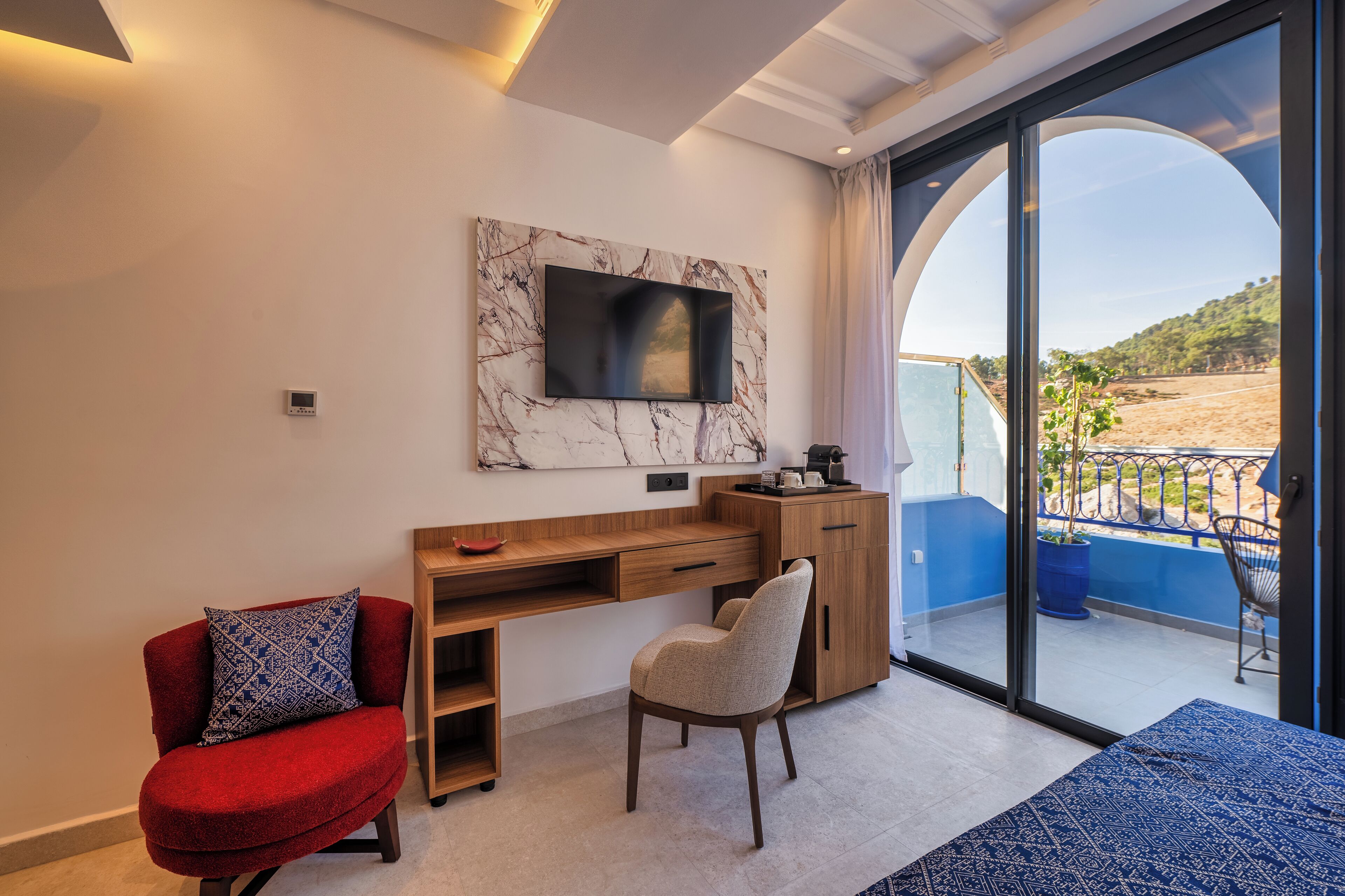 Un dormitorio moderno con un ventanal grande, un televisor instalado en una pared de mármol, un escritorio de madera, un sillón rojo y vista a una playa.