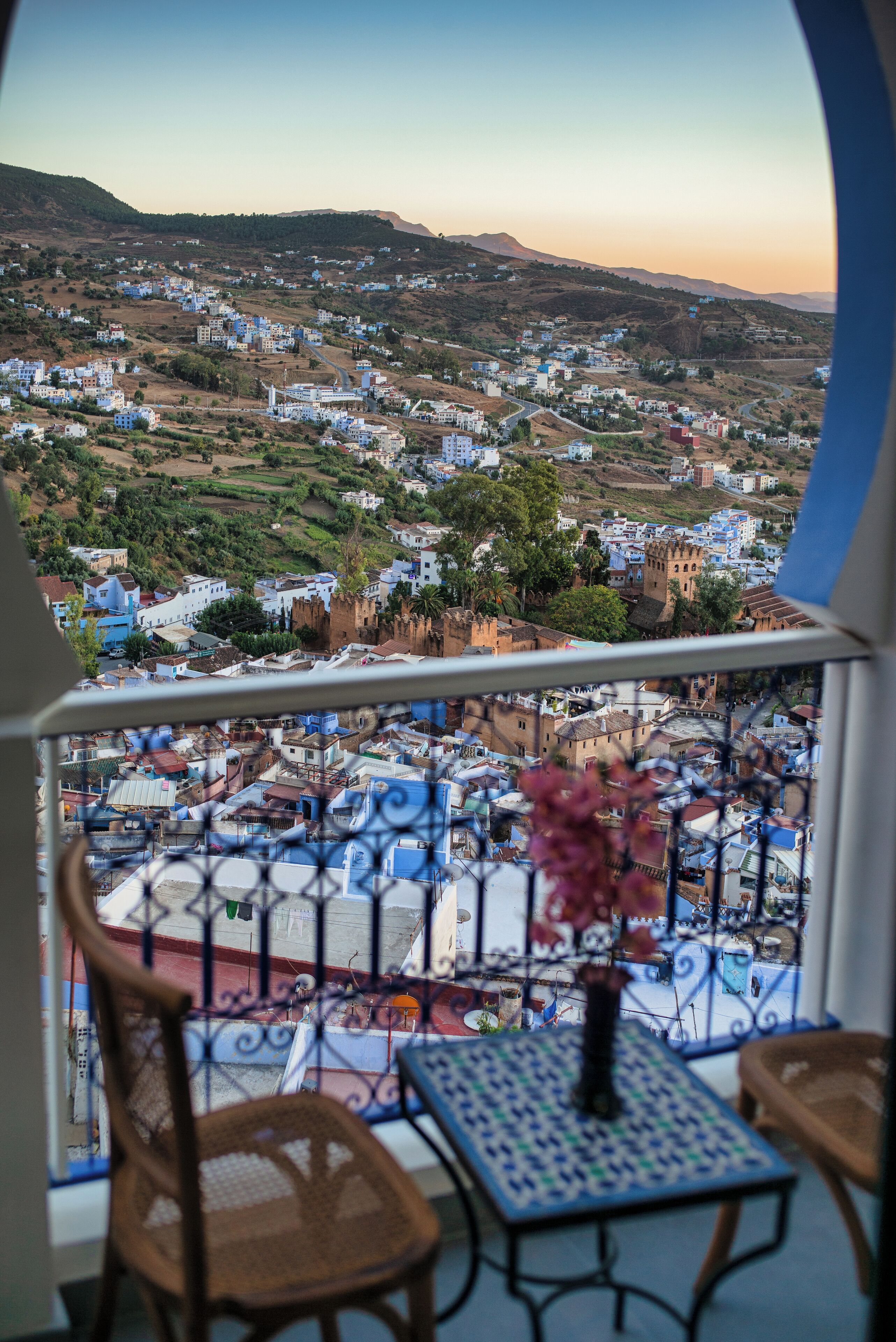 A balcony with a view of a town, a table with a vase, and two chairs.