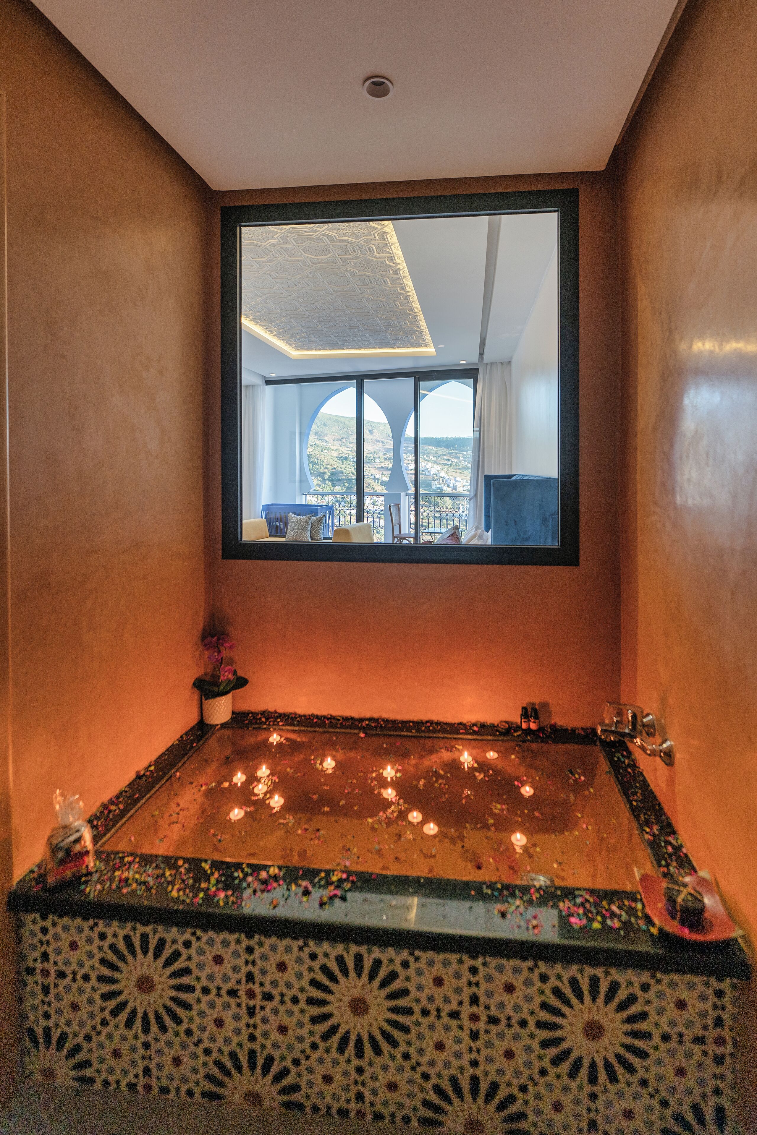 A bathroom with a large bathtub, a mirror above it, and a view of a balcony with a chair and table.