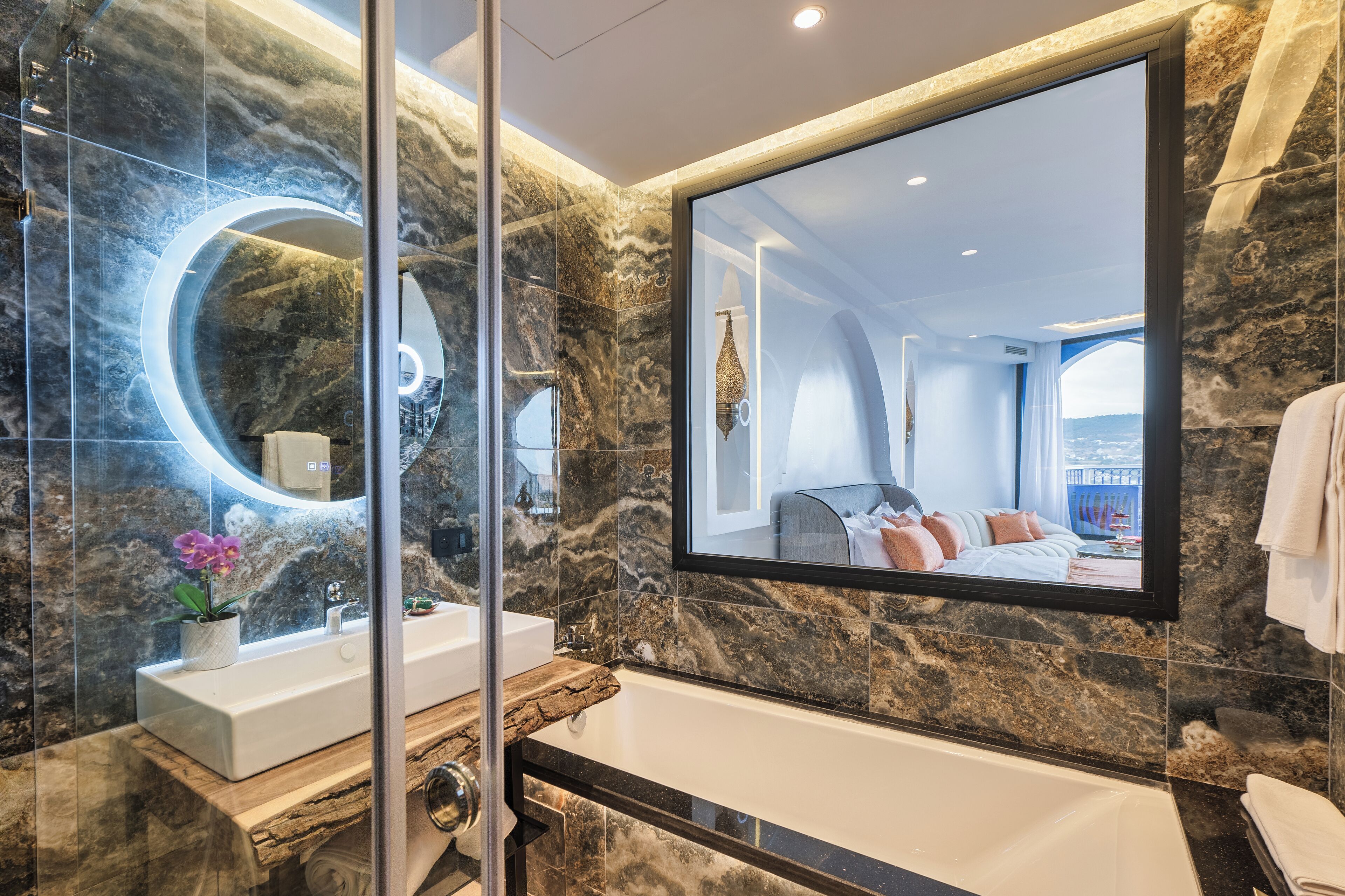 A modern bathroom with a marble wall, a large mirror, a bathtub, and a sink with a potted orchid.