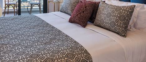 Un dormitorio con una cama grande, ropa de cama estampada, vistas a través de un ventanal y un balcón con sillas.