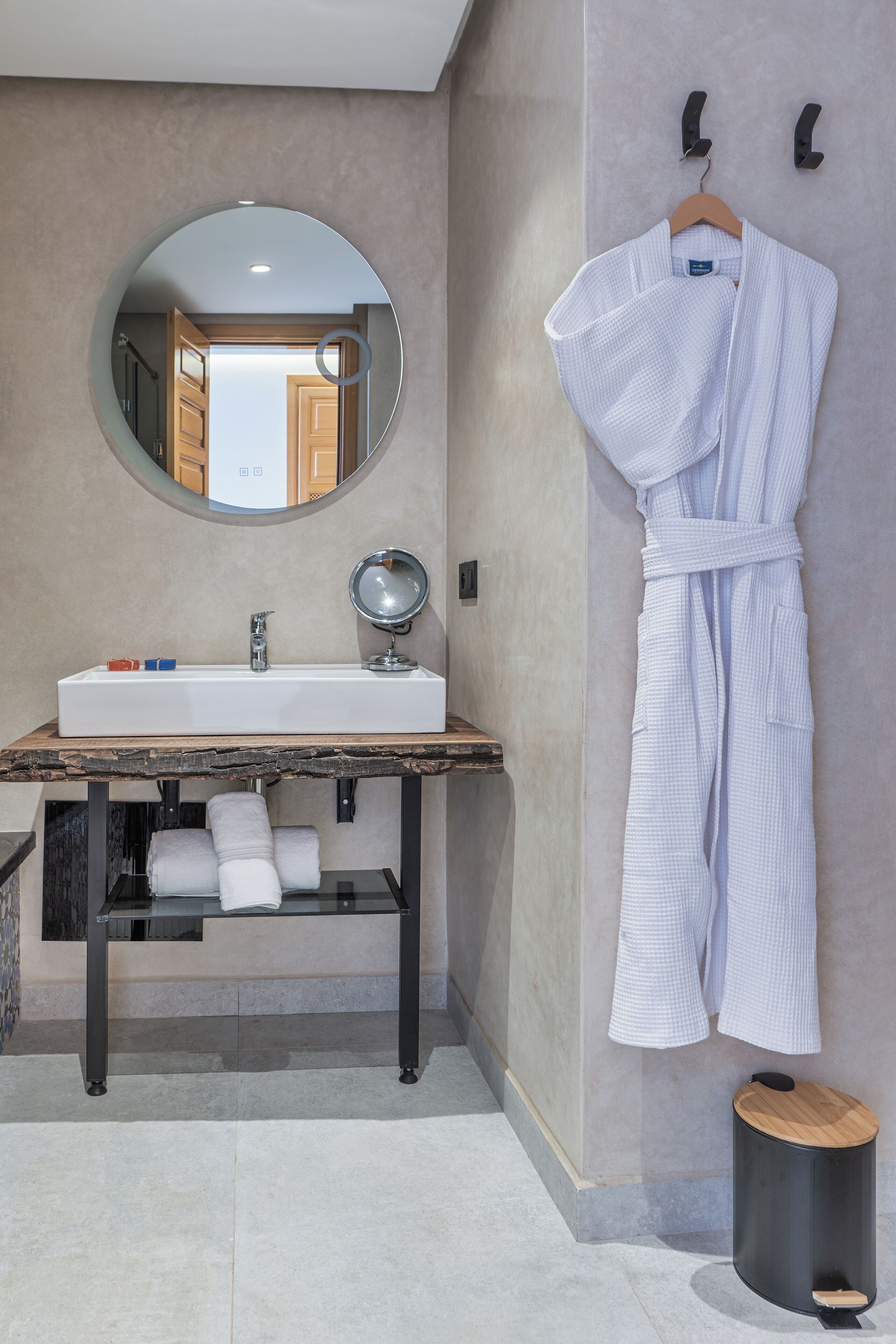 Une salle de bain avec un miroir rond, un lavabo et un meuble-lavabo en bois. Une robe blanche est accrochée au mur.