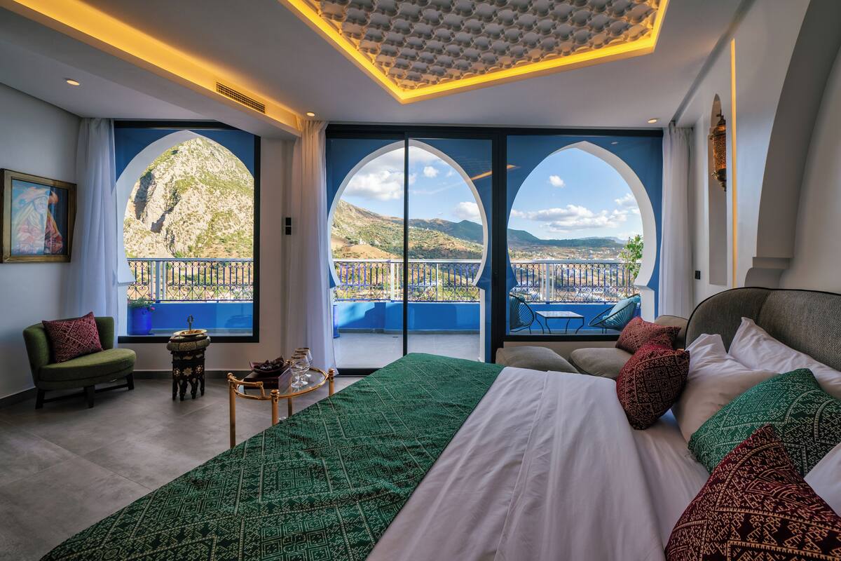 A bedroom with a large bed, two windows with a view of a balcony, a small table with a vase, and a chair.