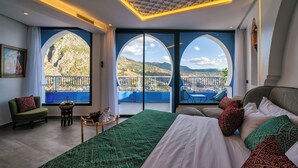 Un dormitorio con una cama grande, dos ventanas que dan a un balcón, una mesita con un florero y una silla.