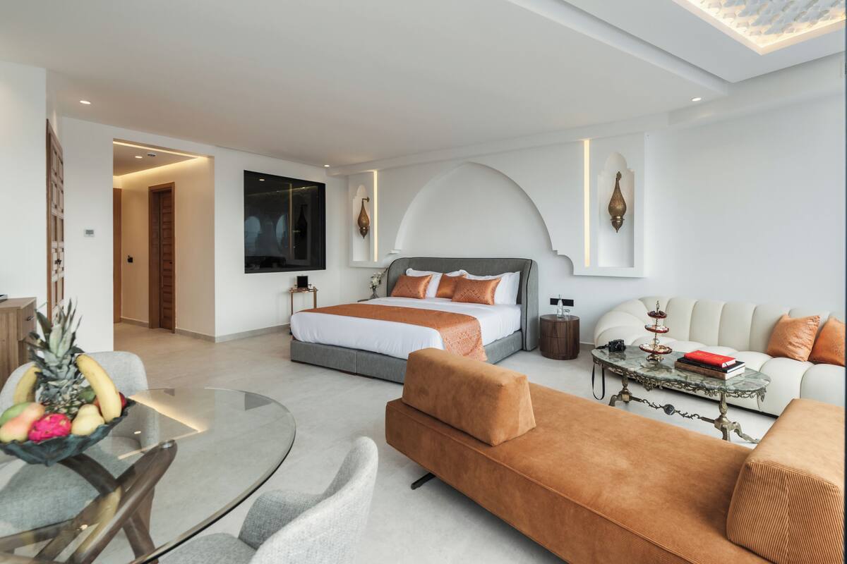 A modern hotel room with a bed, sofa, and a round glass table with fruit.