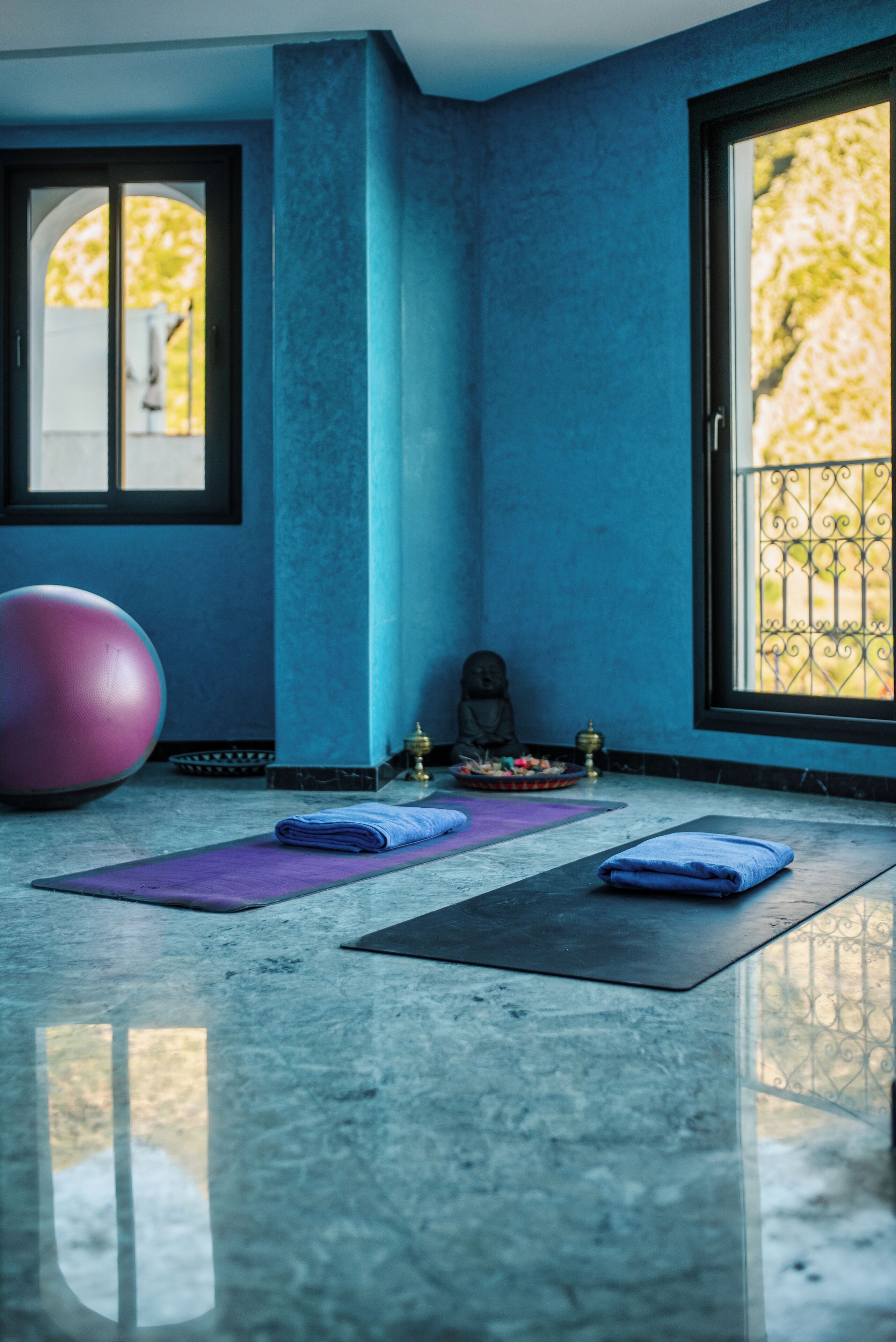 A room with a large exercise ball, two yoga mats, a statue, and a window with a view of greenery.
