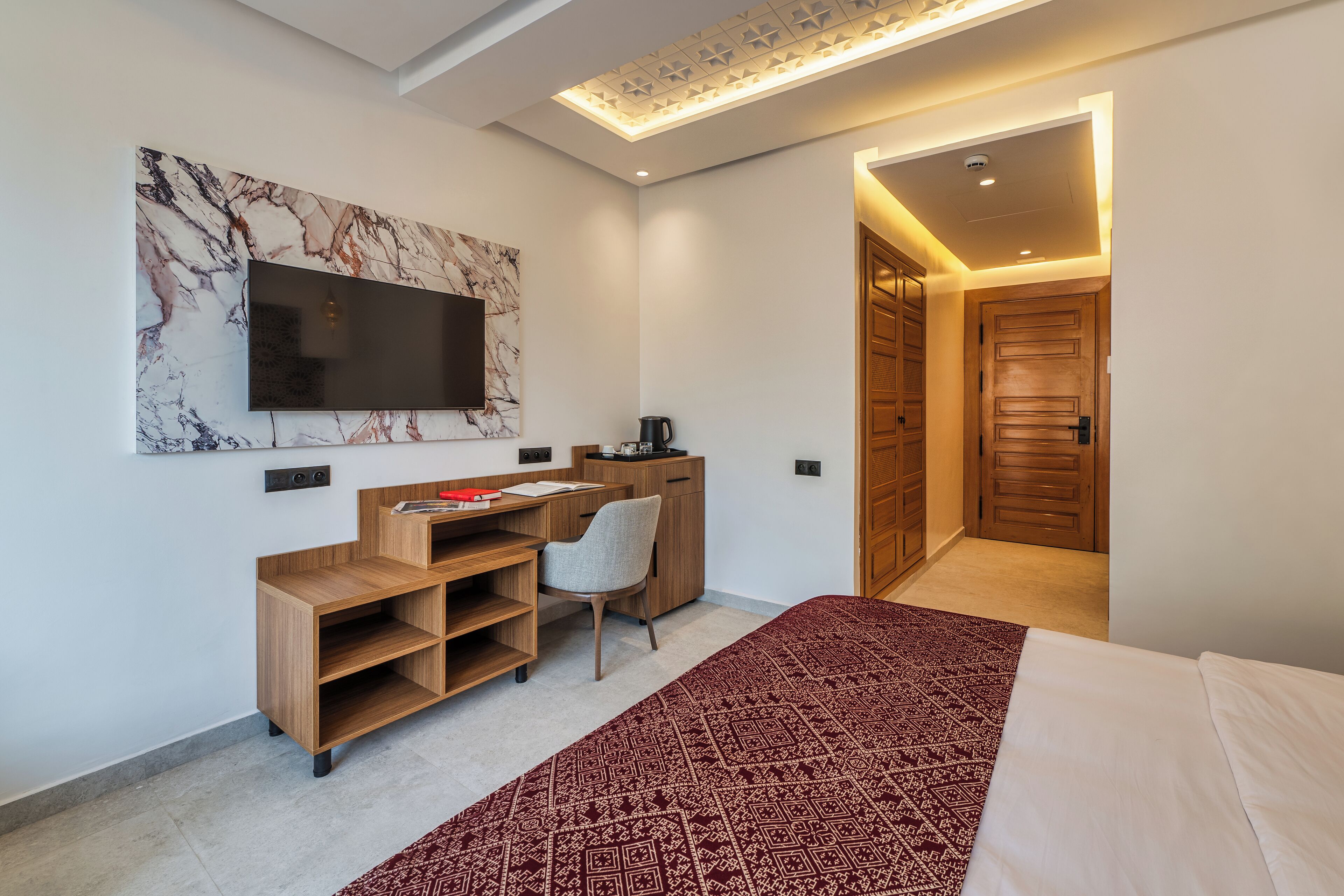 A modern bedroom with a large bed, a desk with a chair, a flat-screen TV mounted on a marble wall, and a wooden wardrobe.