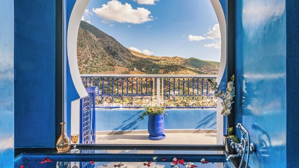 Una habitación con vistas a un paisaje montañoso a través de un amplio ventanal arqueado, un balcón con barandilla y un suelo de baldosas con un diseño geométrico.