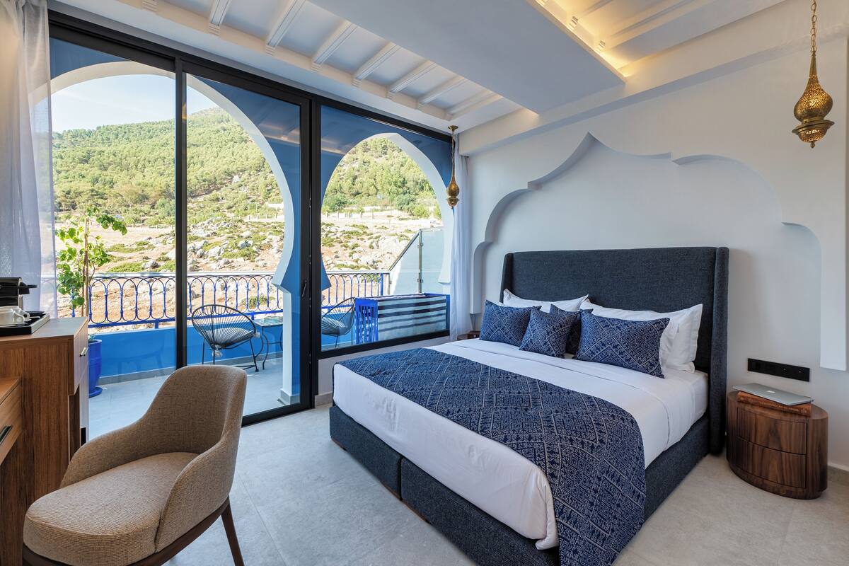 A bedroom with a large bed, a chair, a desk, and a view of a balcony with a railing and a chair.