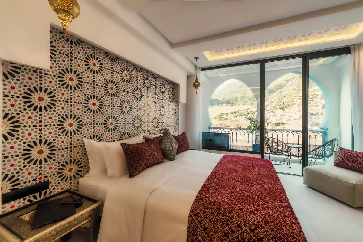 A bedroom with a large bed, a patterned headboard, a balcony with chairs, and a view of the landscape.
