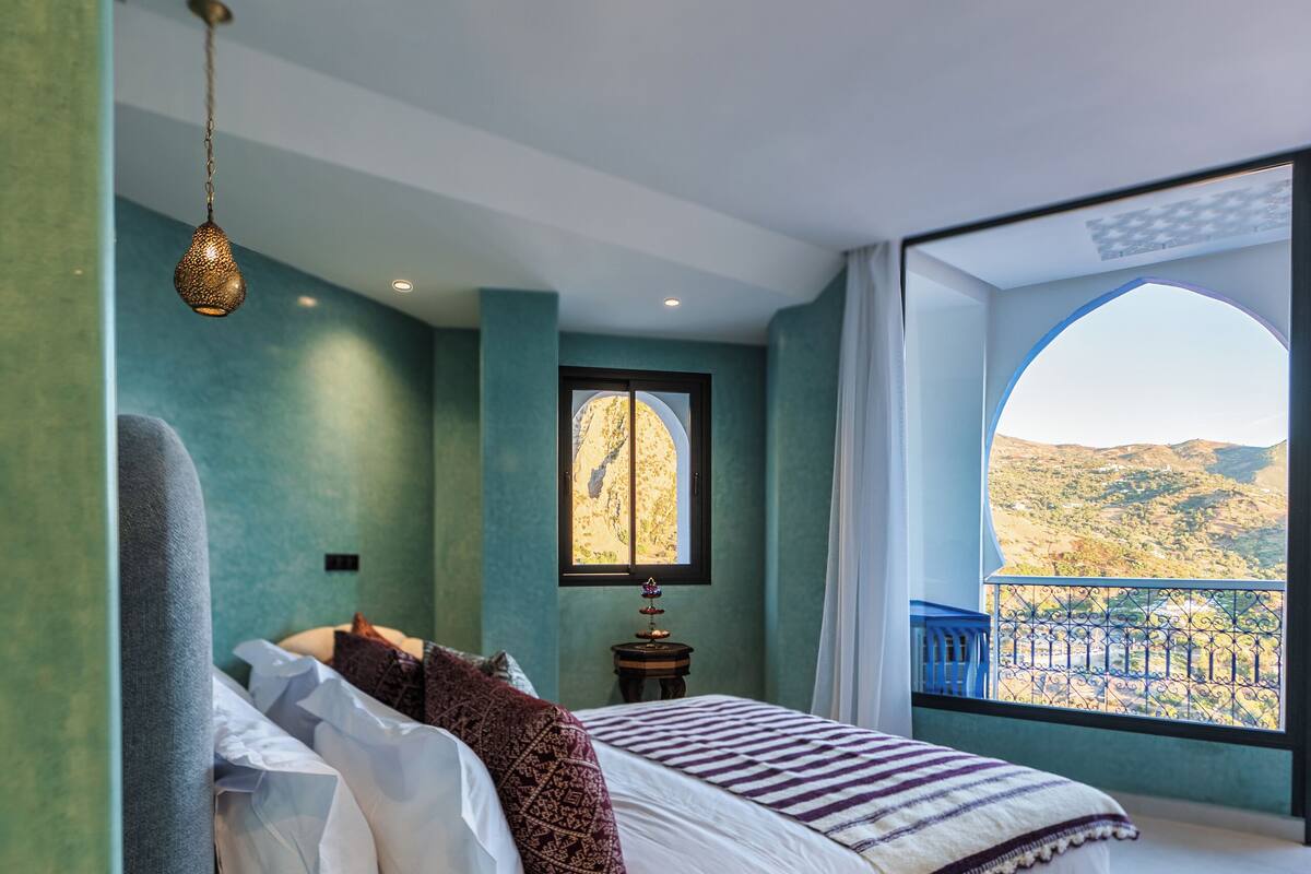 A bedroom with a large bed, a view of the outdoors, and a hanging light fixture.