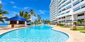 Een zwembad van een resort met helderblauw water, palmbomen en een strand op de achtergrond.